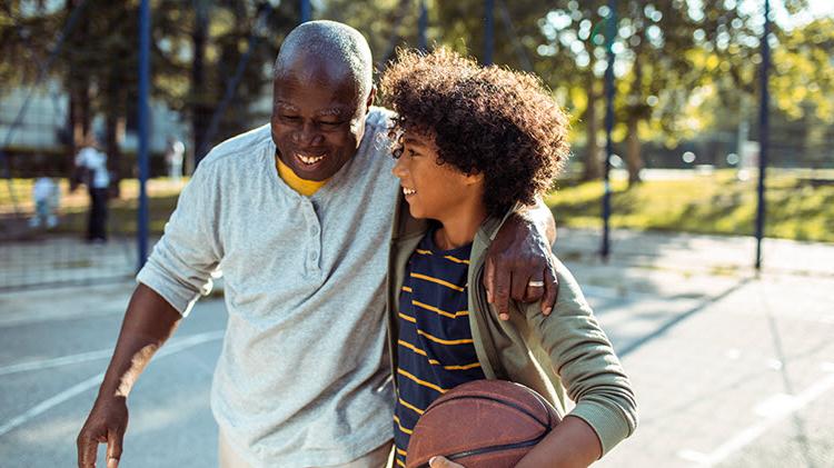 Boy carries a basketball and walks arm-in-arm with his grandfather across a basketball court.